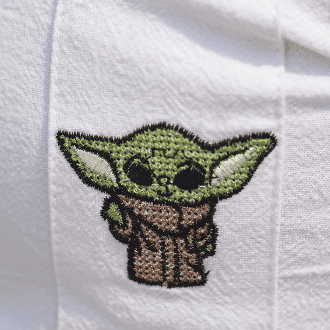 Acercamiento al bordado del pequeño maestro sabio de color verde, símbolo de paz y sabiduría en el universo galáctico, sobre la suave textura de la mezcla lino-algodón.