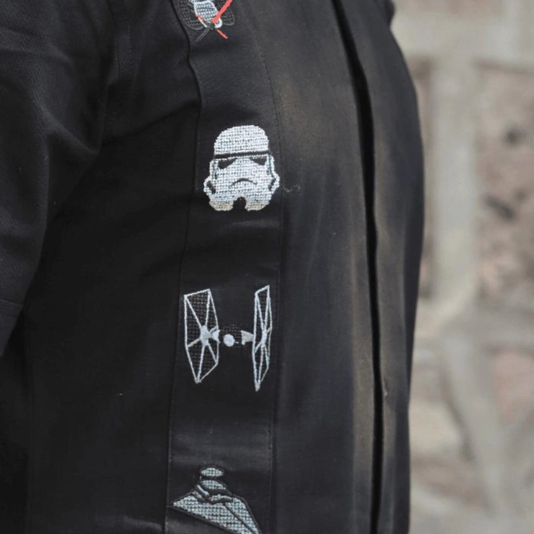 Primer plano del bordado artesanal en punto de cruz con iconos cósmicos y figuras de una galaxia lejana sobre tela negra de algodón premium, resaltando el detalle y la textura.